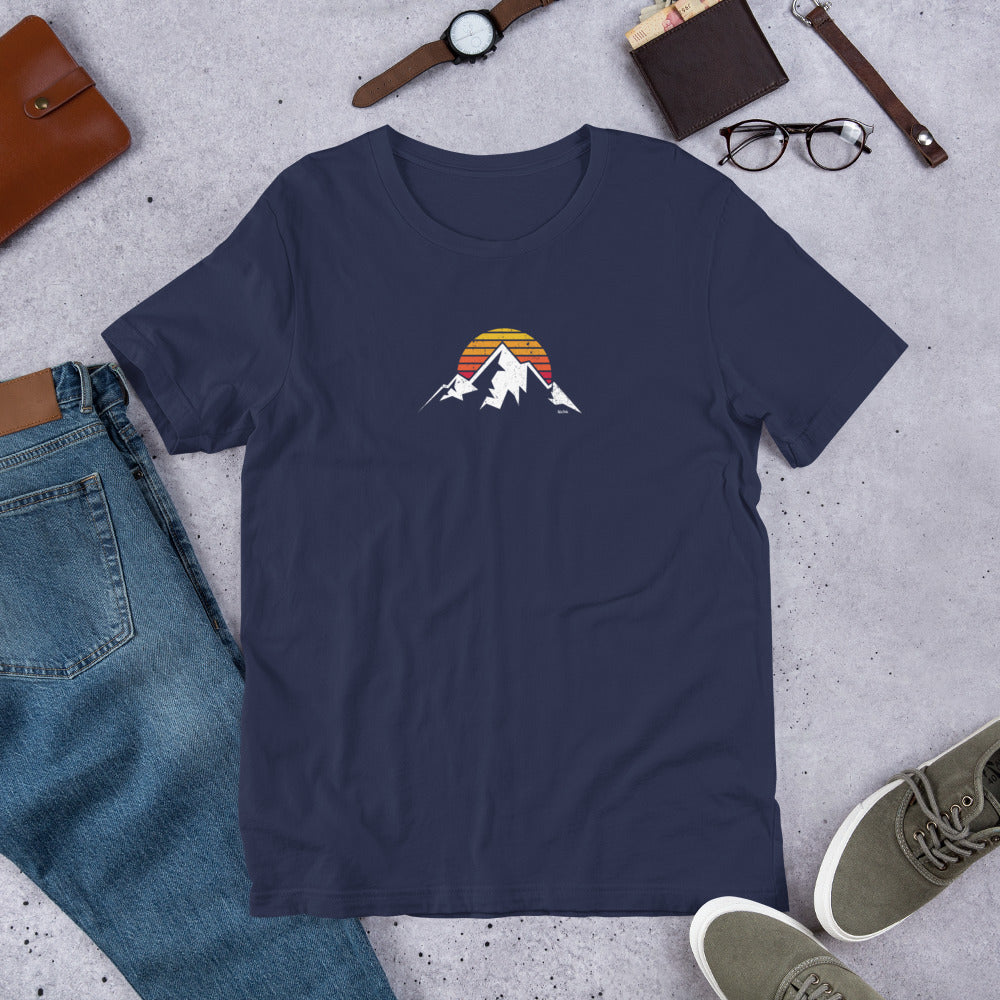 Navy blue t-shirt with mountain and sunset design on a concrete surface with jeans, shoes, and accessories.