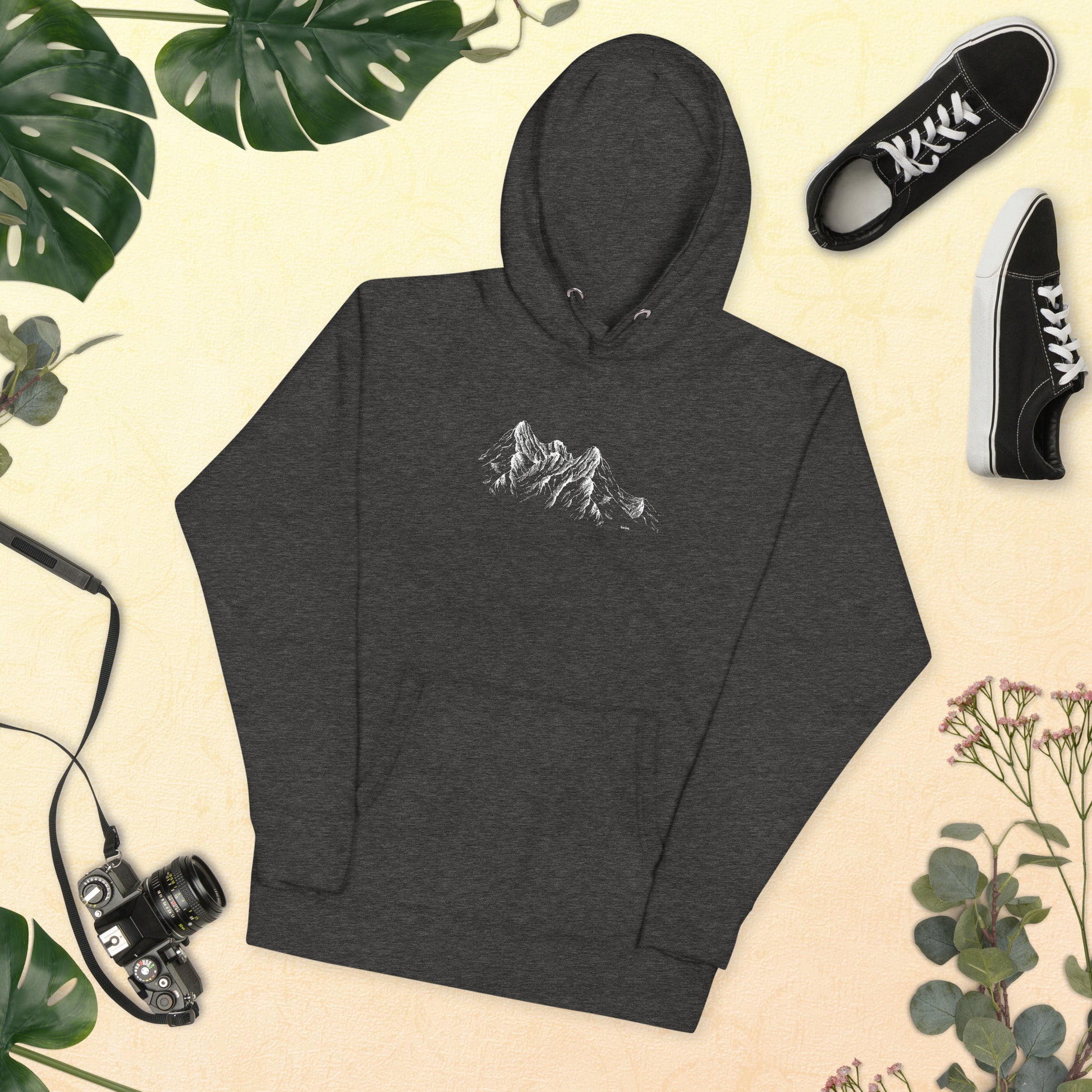 Dark gray hoodie with sketch mountain design on a beige background with plants and shoes.