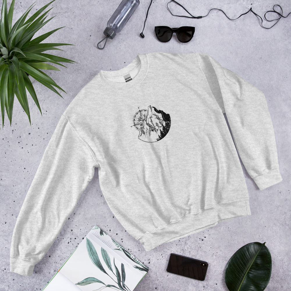 A white sweatshirt with a Mountain Compass graphic design on a gray surface, complete with accessories.