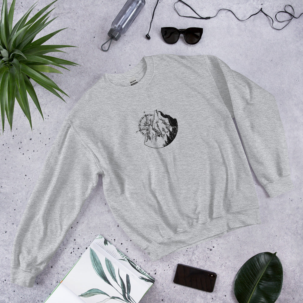 Grey t-shirt with Mountain Compass graphic design on concrete surface with plants and accessories.