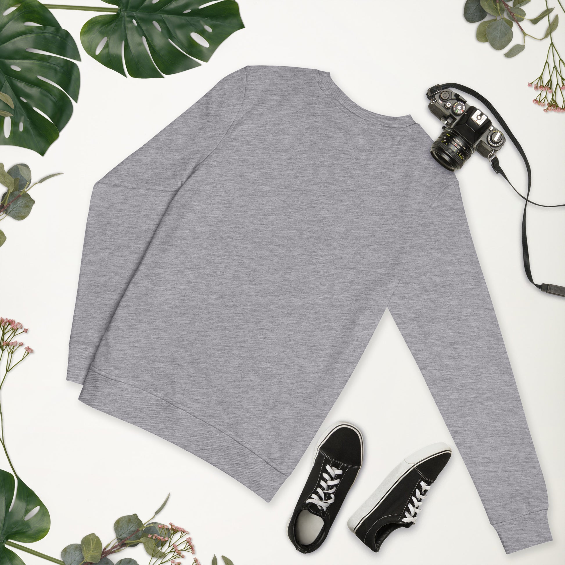 Gray long-sleeve shirt with black sneakers on a white surface surrounded by green leaves.