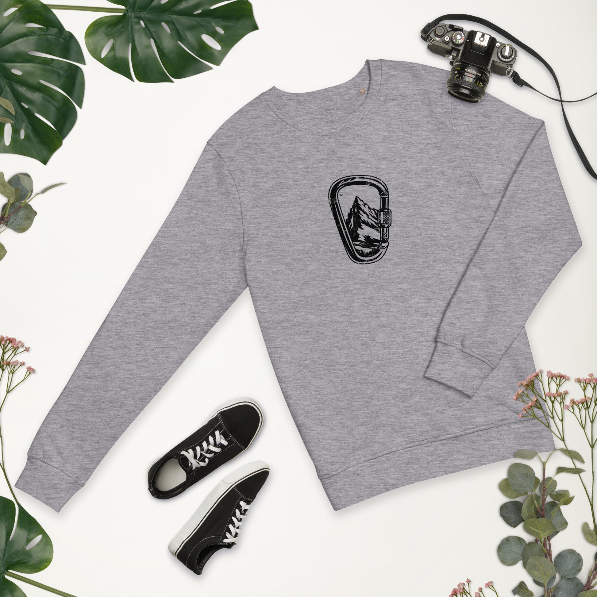 Gray t-shirt with Carabiner Mountain Black design, black sneakers and camera on beige background with green leaves.