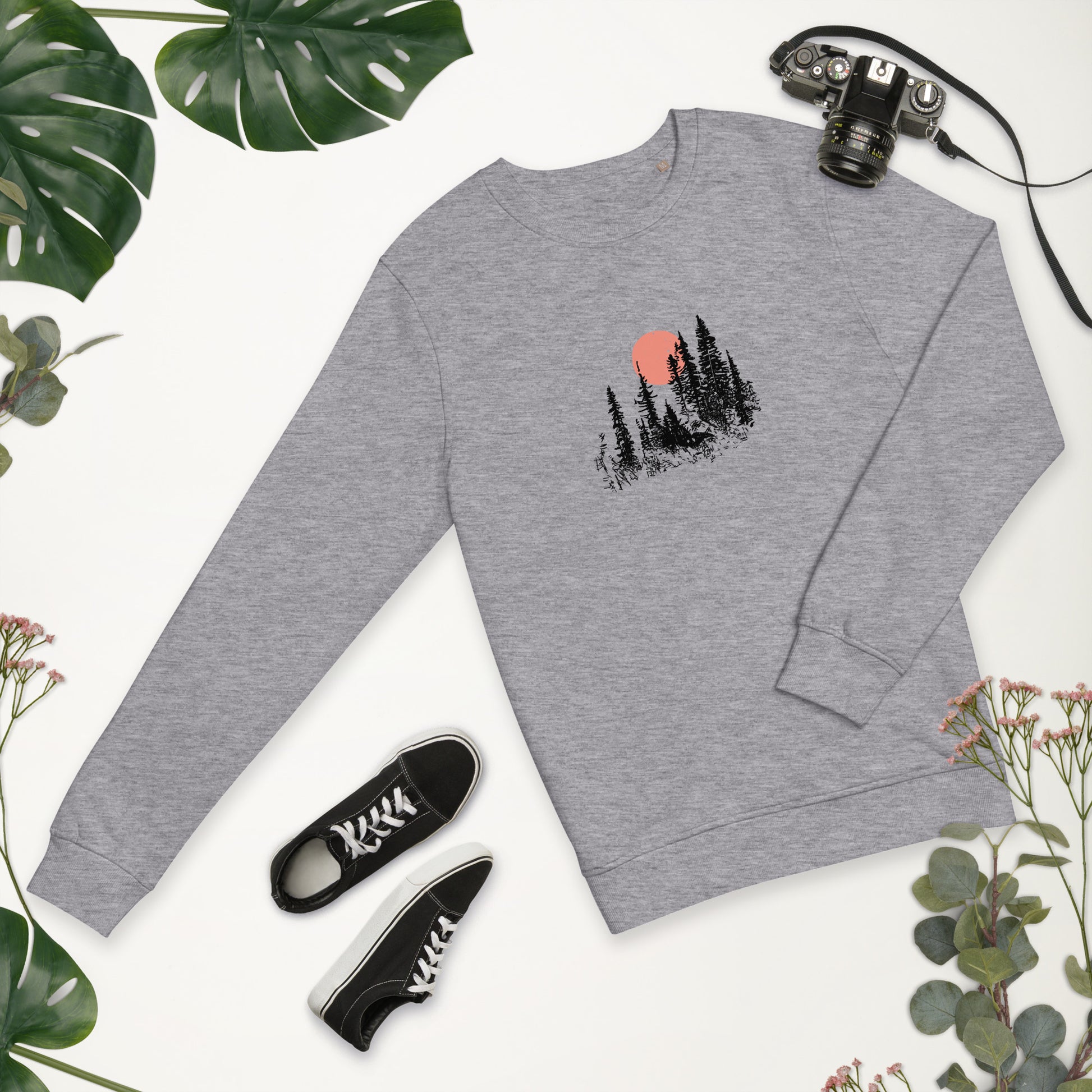 Gray sweatshirt with nature forest graphic, black sneakers, and camera on a white background with greenery.