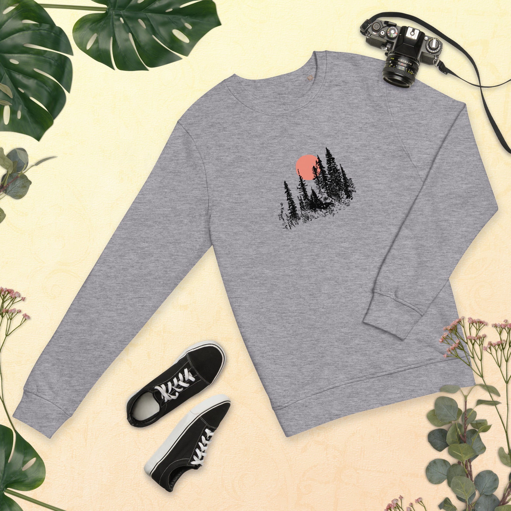 Gray sweatshirt with nature-themed forest design, black sneakers, and camera on a beige background with greenery.