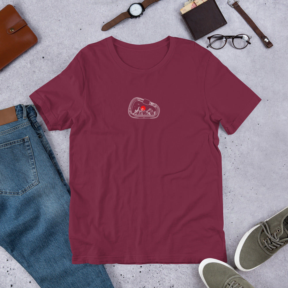 Maroon t-shirt with a graphic design on a gray surface with jeans, shoes, and accessories.